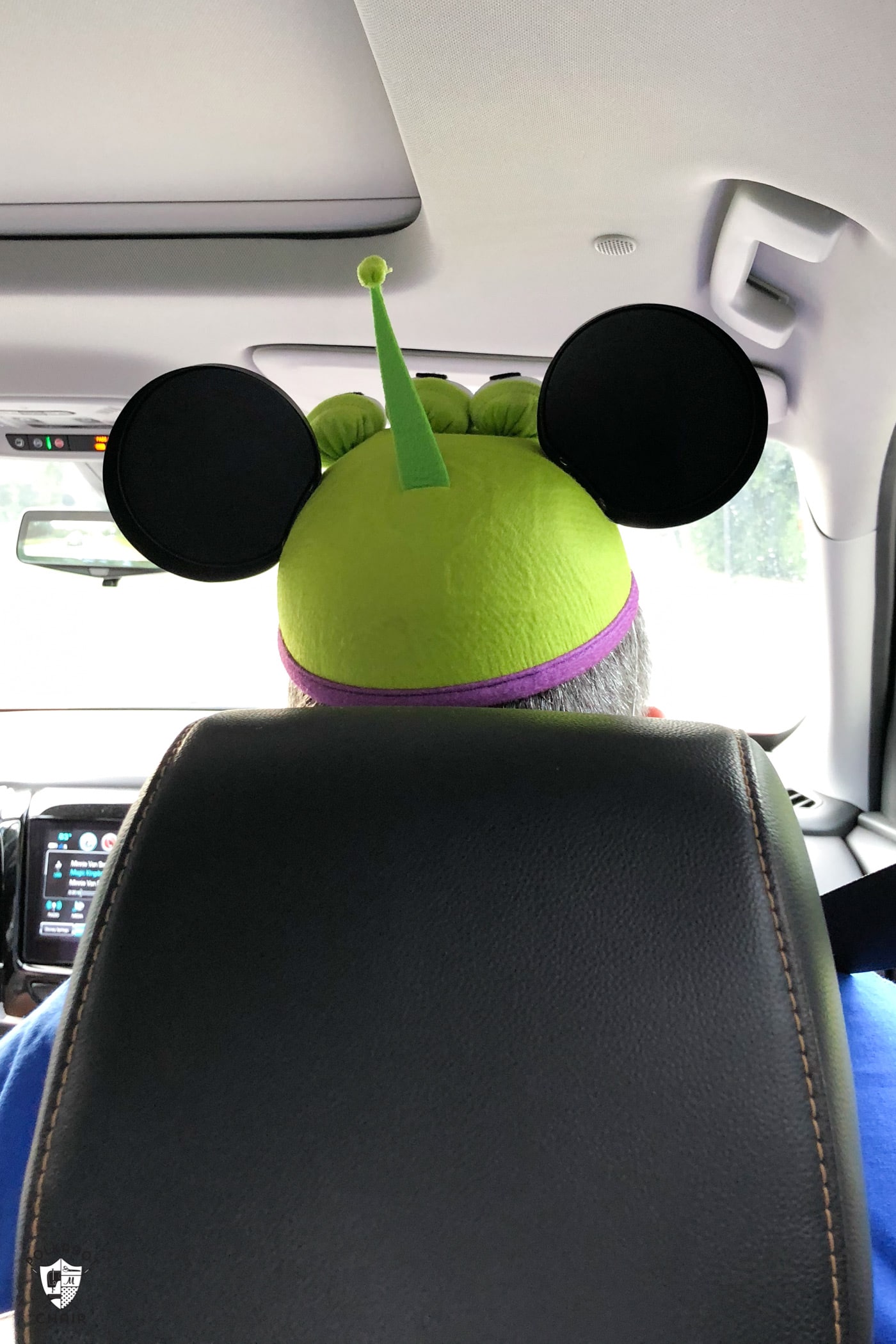 Assis dans une voiture prise par derrière, vous voyez le dossier du siège et le haut de la tête d'un homme portant un chapeau vert à oreilles de Mickey extraterrestre.